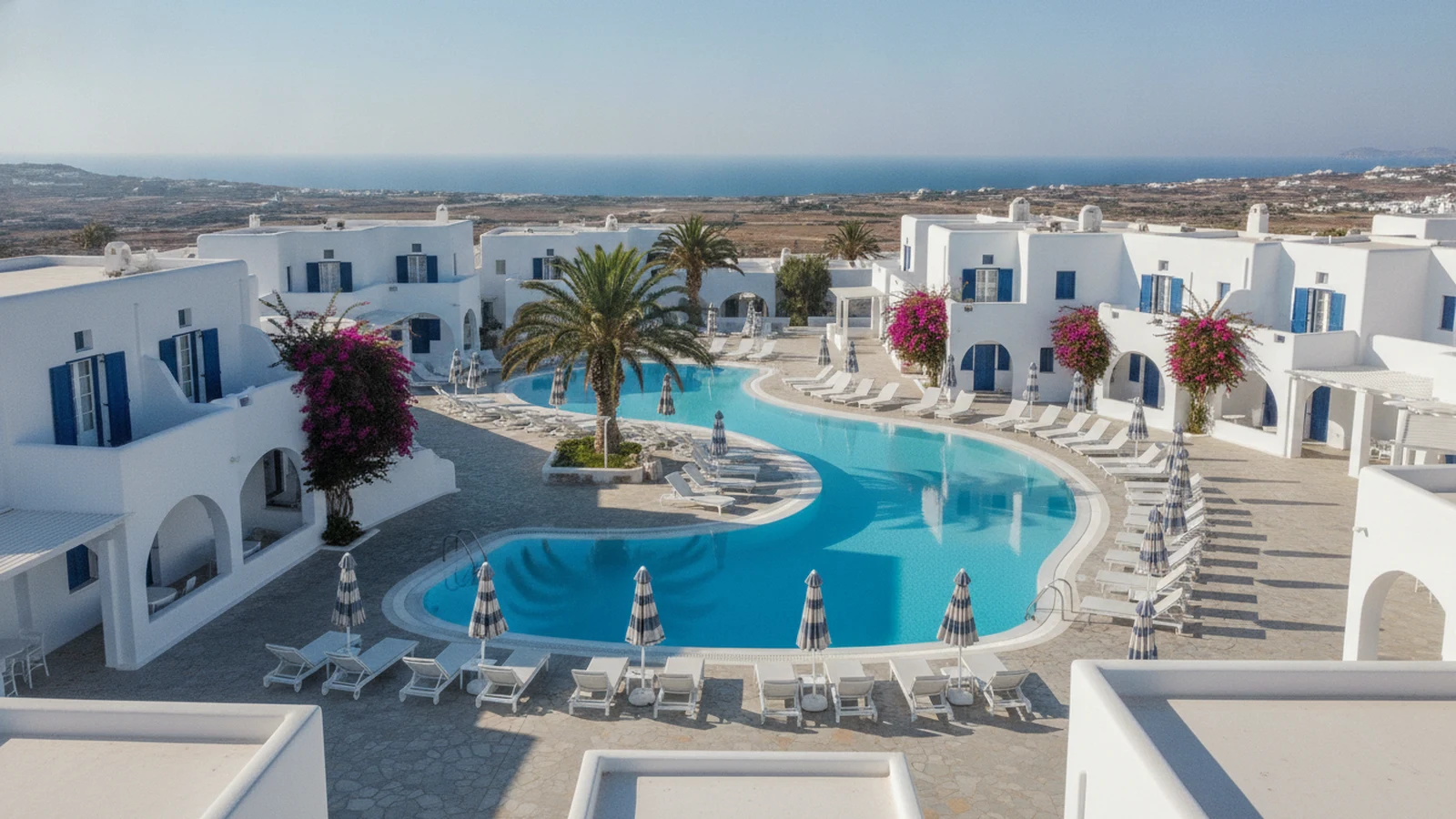 Bílé kykladské budovy resortu s bazény a lehátky ve Fiře na Santorini, v dálce moře