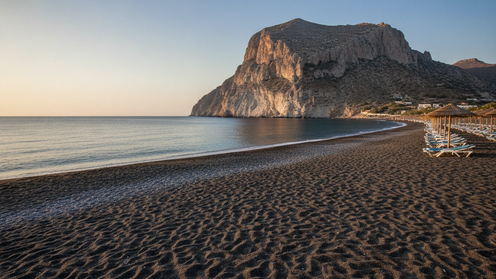 Černý sopečný písek na pláži Perissa na Santorini se skalním masivem Mesa Vouno v pozadí